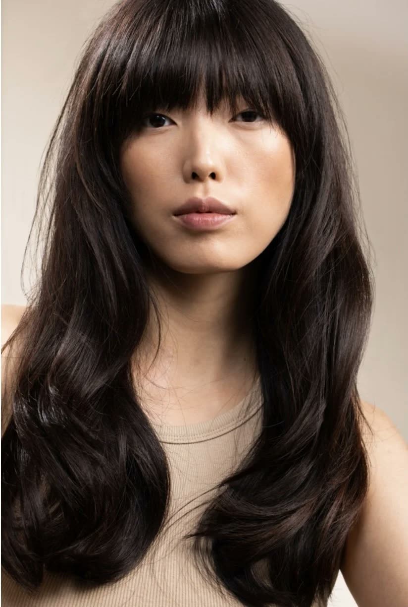 Woman with long, glossy dark hair and full bangs in a beige top