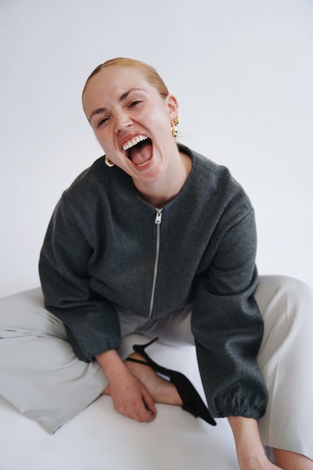 Chloie Costello laughs, seated in a grey zip bomber, cream trousers and black slingbacks