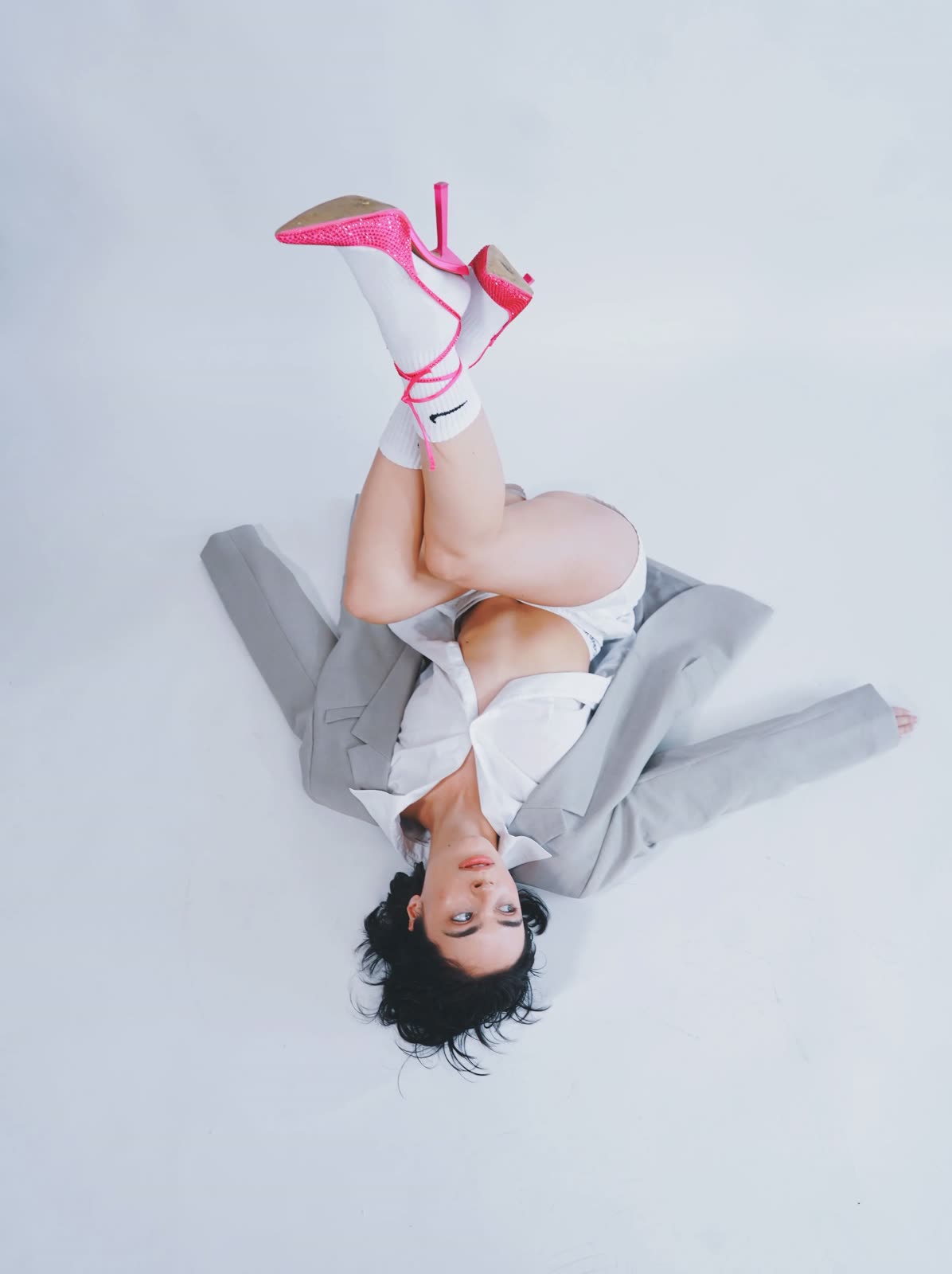 Reilly Hail lies upside down on a white floor in a grey blazer, white socks and pink heels
