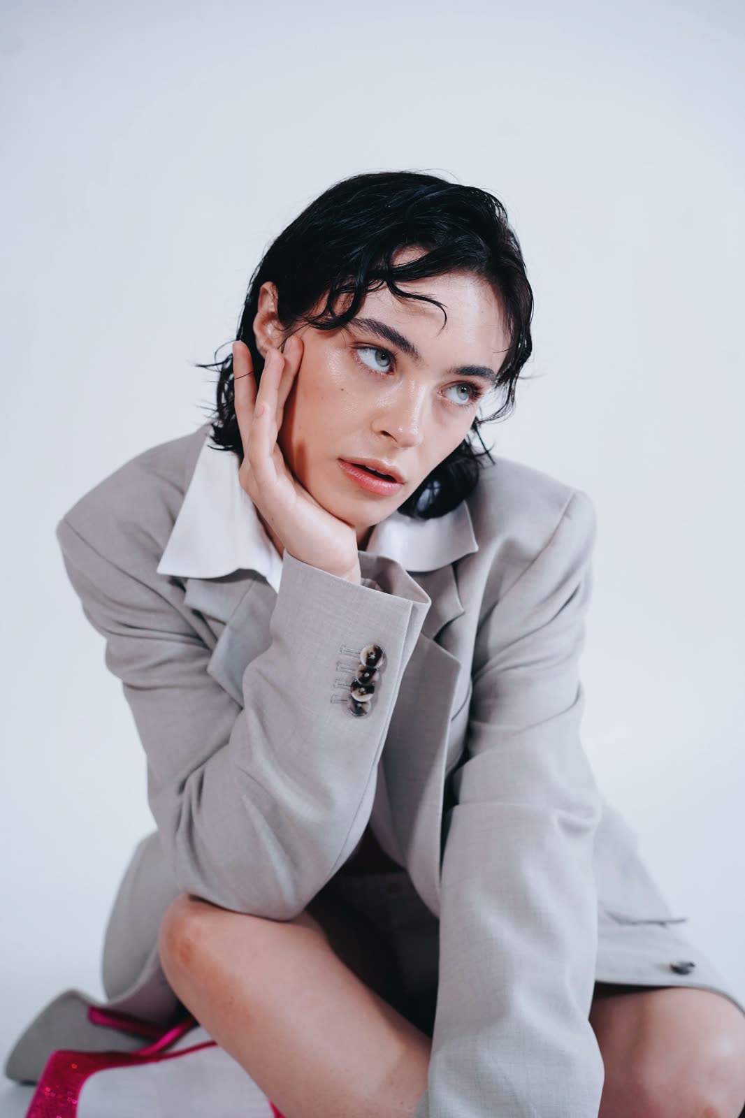 Reilly Hail sits in a grey blazer with a white collar, hand on cheek, pink heels