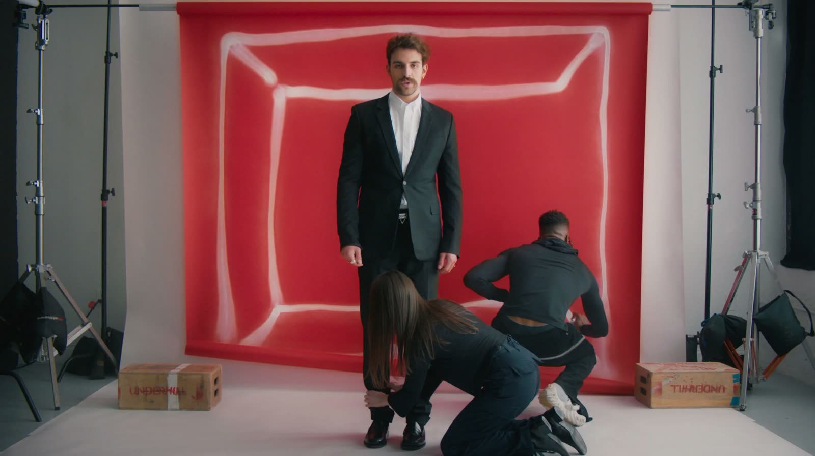 Two crew members kneel to fit a man's black suit trousers before a red backdrop, Facetime video still