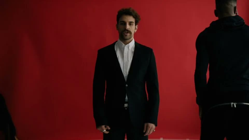 Man in a black suit and white shirt in front of a red backdrop, Facetime video still