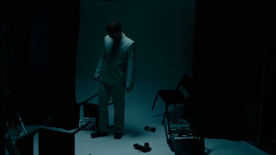 Man in a loose pale suit in a dim teal-lit studio among folding chairs, Facetime video still