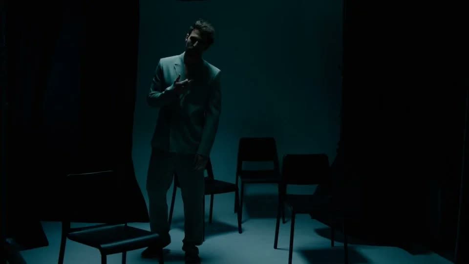 Man in a pale suit adjusts his jacket in a dark teal-lit studio, Facetime video still