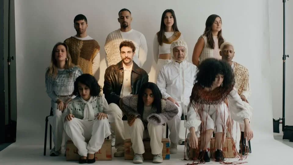 Group in crochet and knit looks on a white set, a man in a brown leather jacket at center, Facetime video still