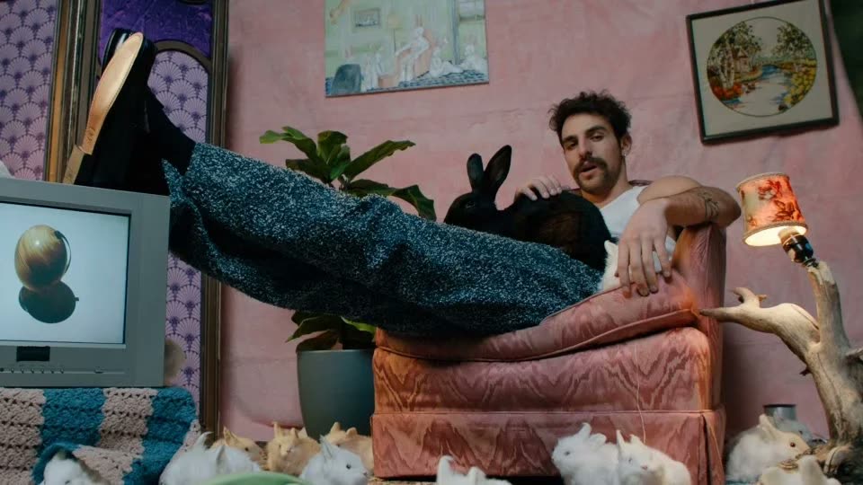 Man lounges on a pink armchair in speckled knit trousers, a black rabbit on his chest, Facetime video still
