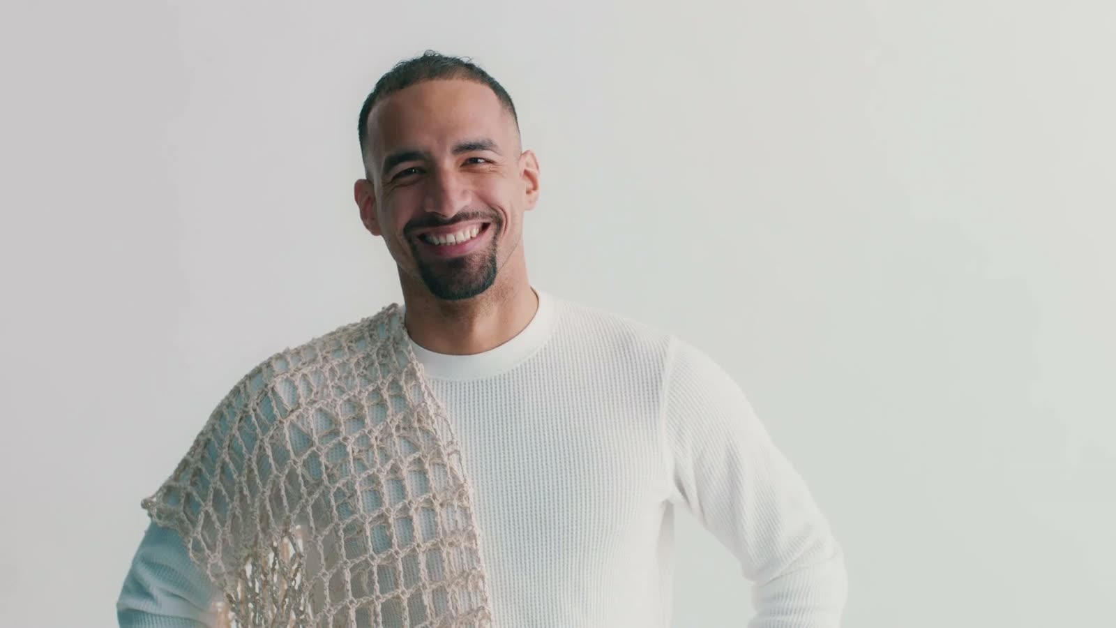 Smiling man in a white knit top with a crochet mesh panel over one shoulder, Facetime video still