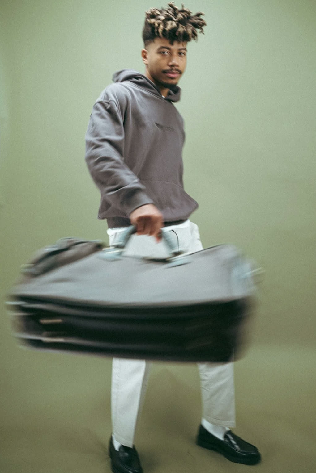 Man with bleached twists in a charcoal hoodie, white trousers and loafers swings a duffel bag