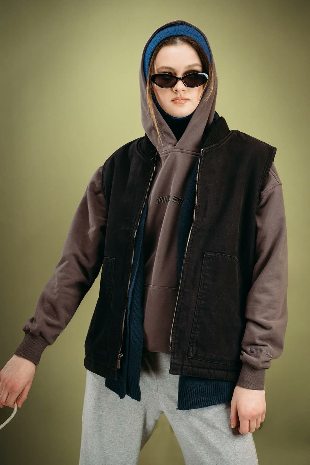 Woman in a brown hoodie under a black canvas vest, blue beanie, sunglasses and grey sweatpants