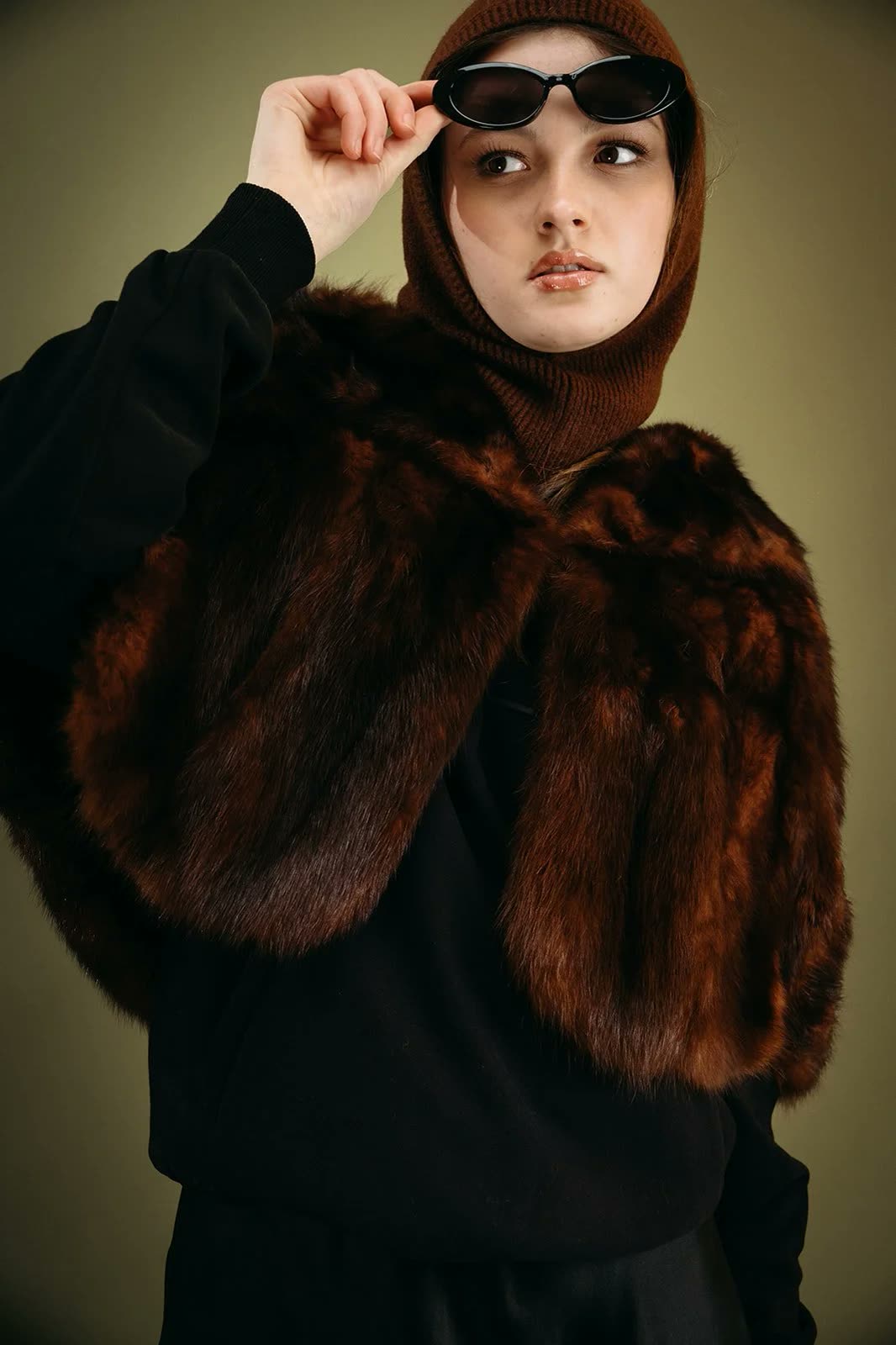 Woman in a brown balaclava and fur stole over a black hoodie lowers her sunglasses