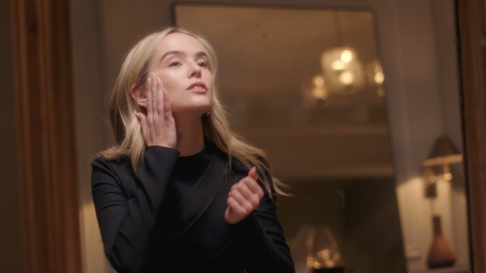 Zoey Deutch in a black long-sleeve dress touches her face at a mirror, Estée Lauder film still
