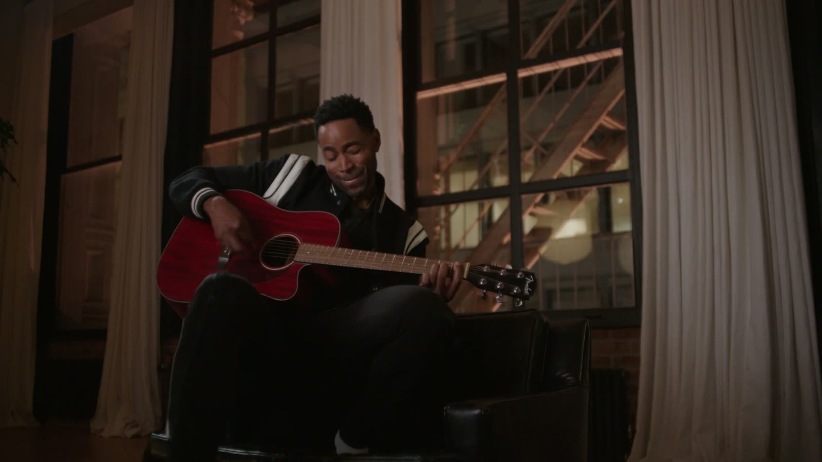 Jay Ellis in a black varsity jacket plays a red guitar by a loft window, Estée Lauder film still
